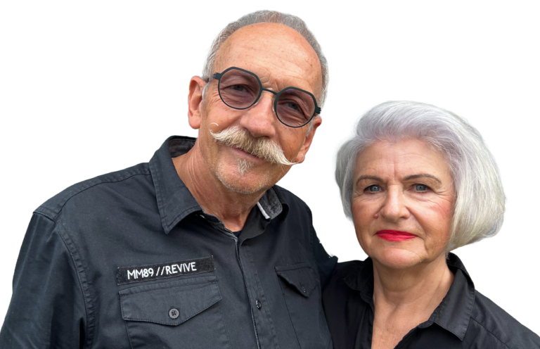 Ein Porträt eines älteren Paares vor einem schwarzen Hintergrund. Der Mann trägt eine Brille, hat einen Schnurrbart und lächelt sanft, während er ein schwarzes Hemd mit der Aufschrift 'MM89 // REVIVE' trägt. Die Frau neben ihm hat weißes Haar, trägt roten Lippenstift und ein passendes schwarzes Hemd, und blickt ernst in die Kamera.
